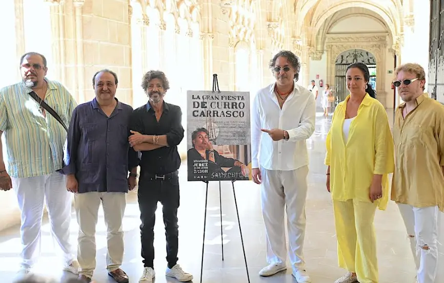 Six people stand in a sunlit arched hallway beside a festival poster on an easel, promoting La Gran Fiesta de Curro Carrasco.