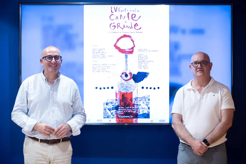 Two men stand on either side of a large white poster mounted on a blue wall, smiling at the camera.