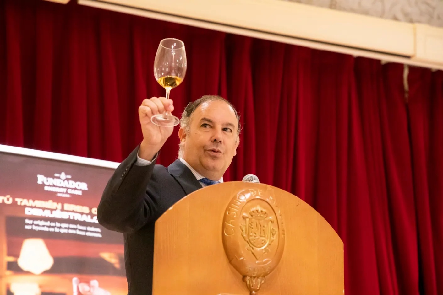 Man in a suit raises a wine glass in a toast from a wooden podium with a crest, red stage curtains behind