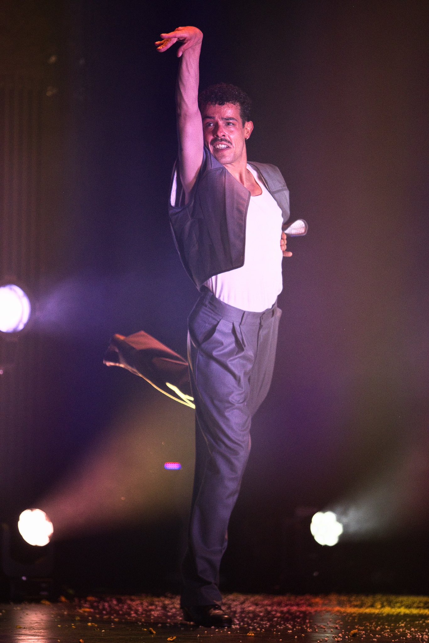 Male performer on stage with raised right arm, wearing a white shirt and gray jacket, under dramatic stage lighting.