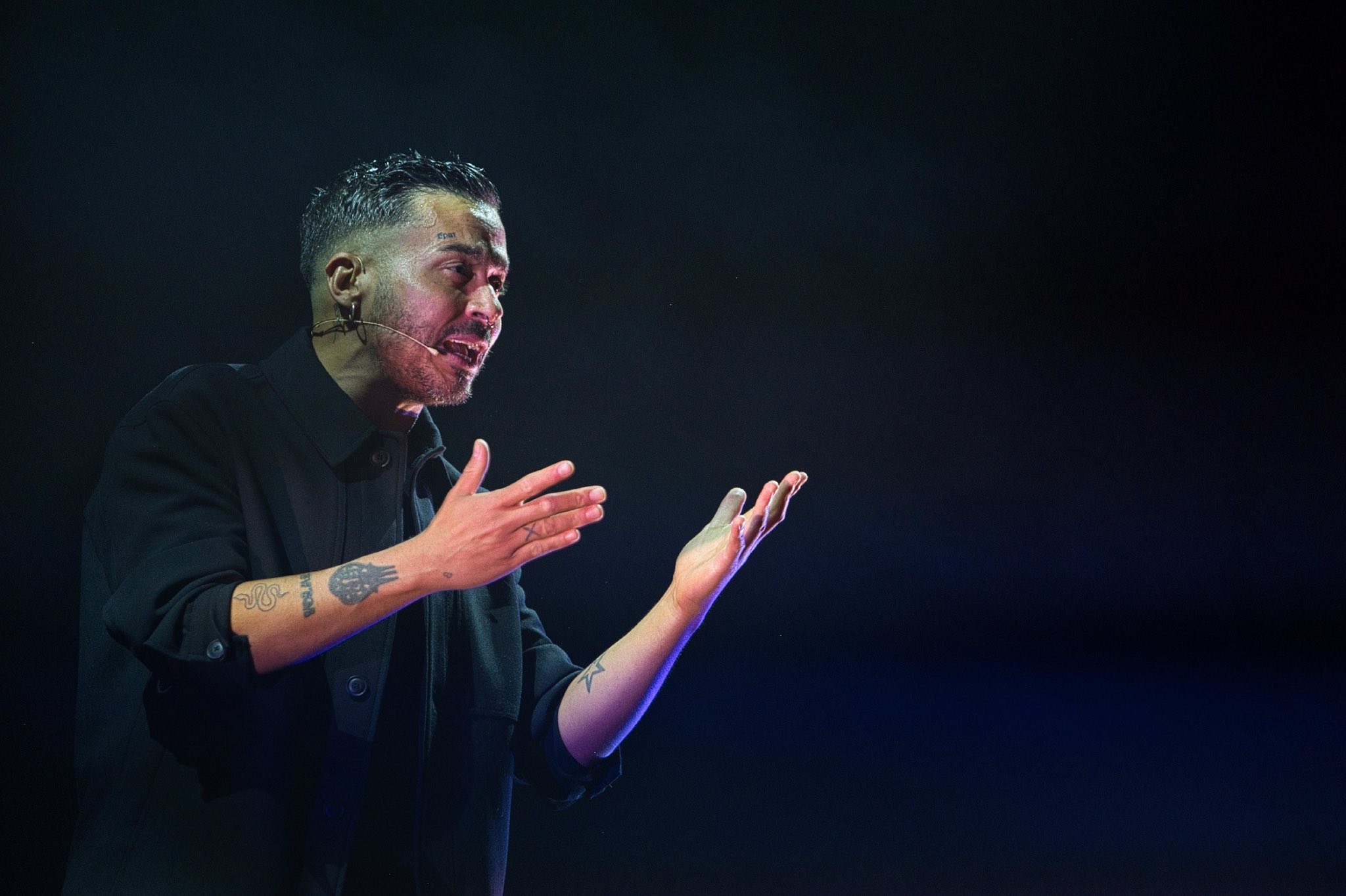 Male singer on stage with a headset mic, wearing a dark jacket, gesturing with hands against a dark background on a night concert.