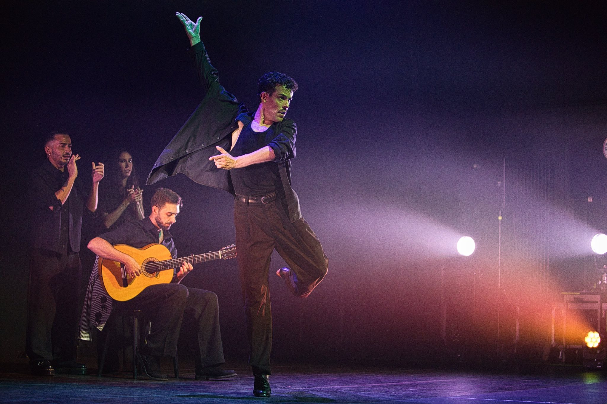Male dancer mid-leap on stage with right arm raised, accompanied by a guitarist and backup singers on a dark, smoky concert set.