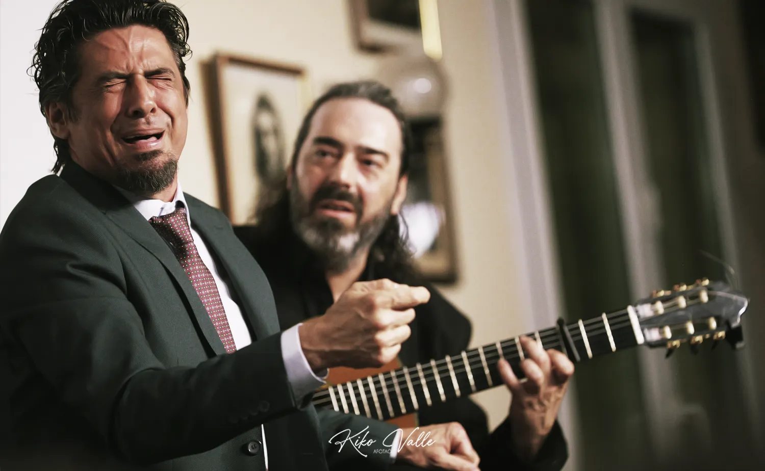 Two men in a room: one in a suit crying or moved, while the other with a beard plays an acoustic guitar beside him, creating an emotional moment.