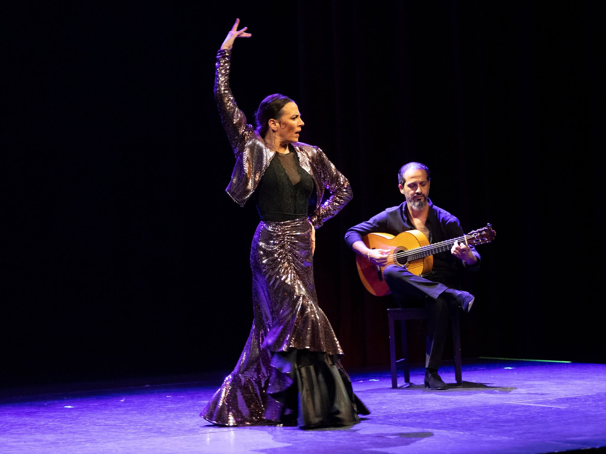 Female flamenco dancer in a shiny sequined gown raises one arm on a dark stage, guitarist seated behind her