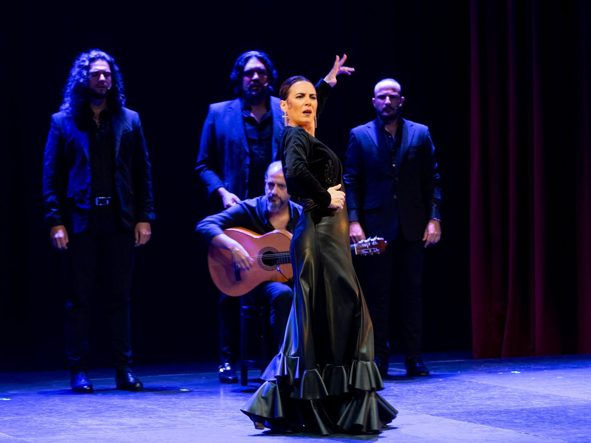 Center-stage flamenco dancer in a long black dress with ruffles, performing on a dim blue-lit stage as four male guitarists stand behind her in dark suits. (center-stage dancer with accompaniment)