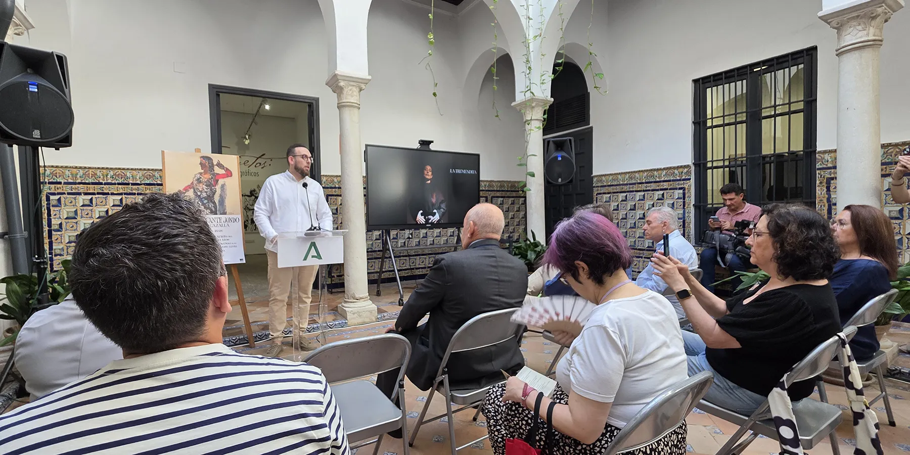 Speaker at a white podium addressing a small audience in a sunlit tiled courtyard.