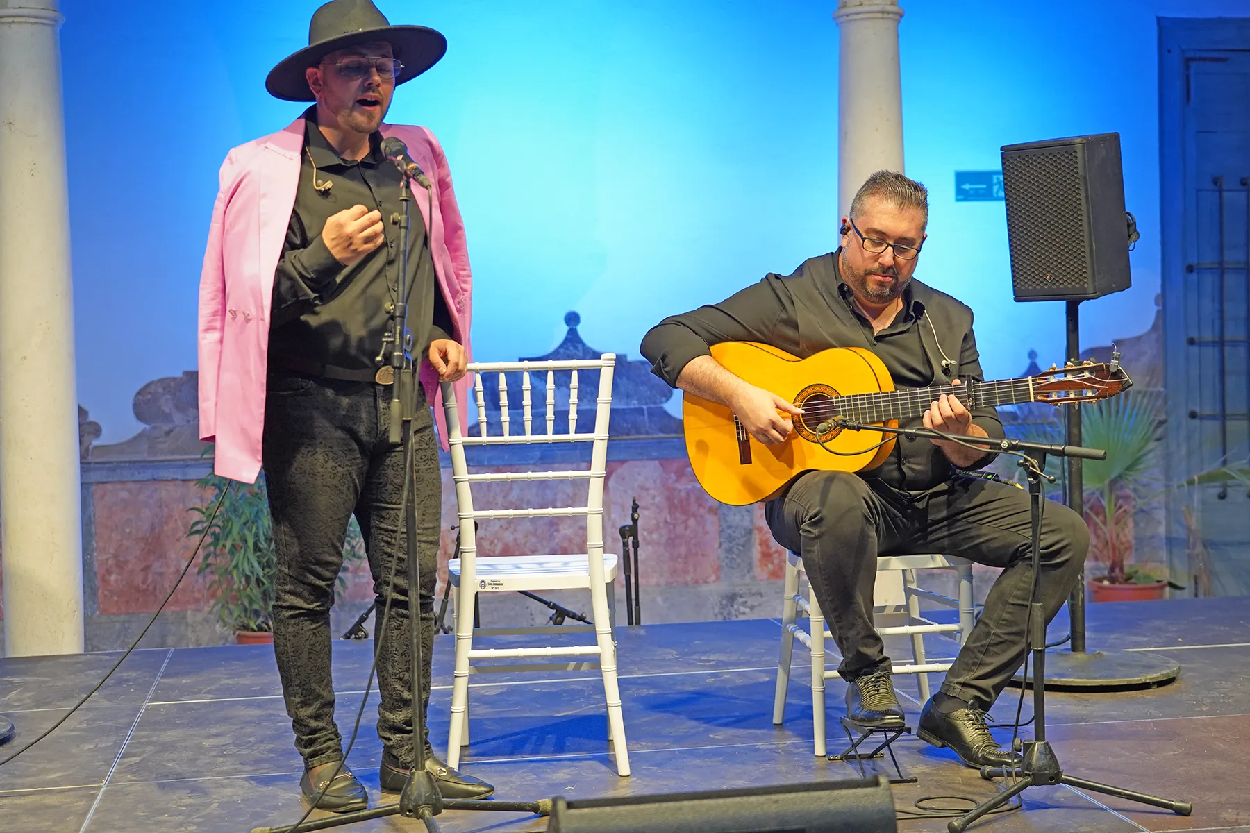 Singer in a pink blazer and hat performs into a microphone while a guitarist in black plays an orange classical guitar on a blue-lit stage.