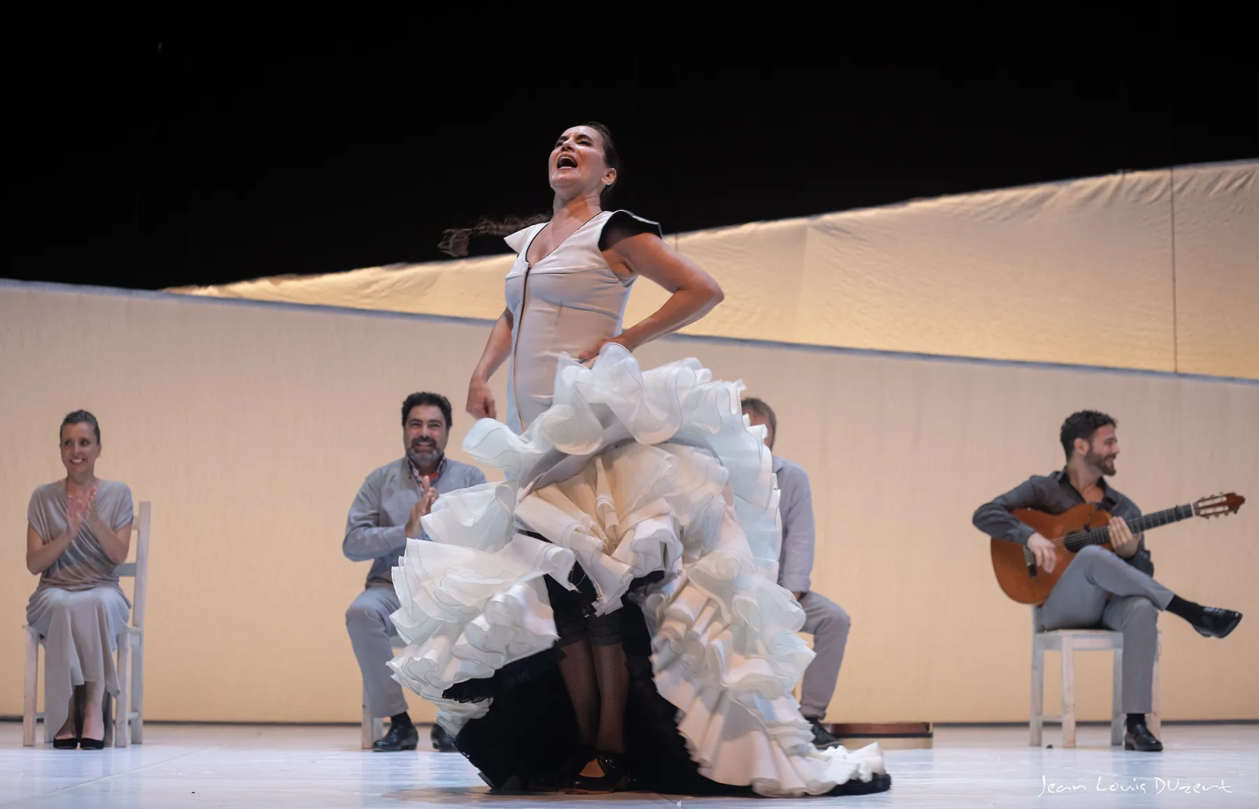 Dancer in a voluminous white ruffled gown performing on stage as teammates clap and a guitarist plays nearby.