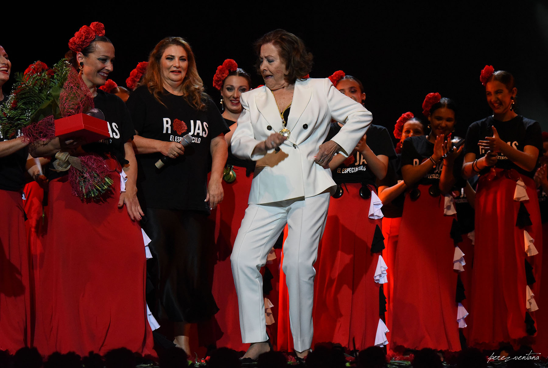 A woman in a white suit dances center stage while fellow performers in black tops and red skirts applaud and hold a bouquet of flowers.