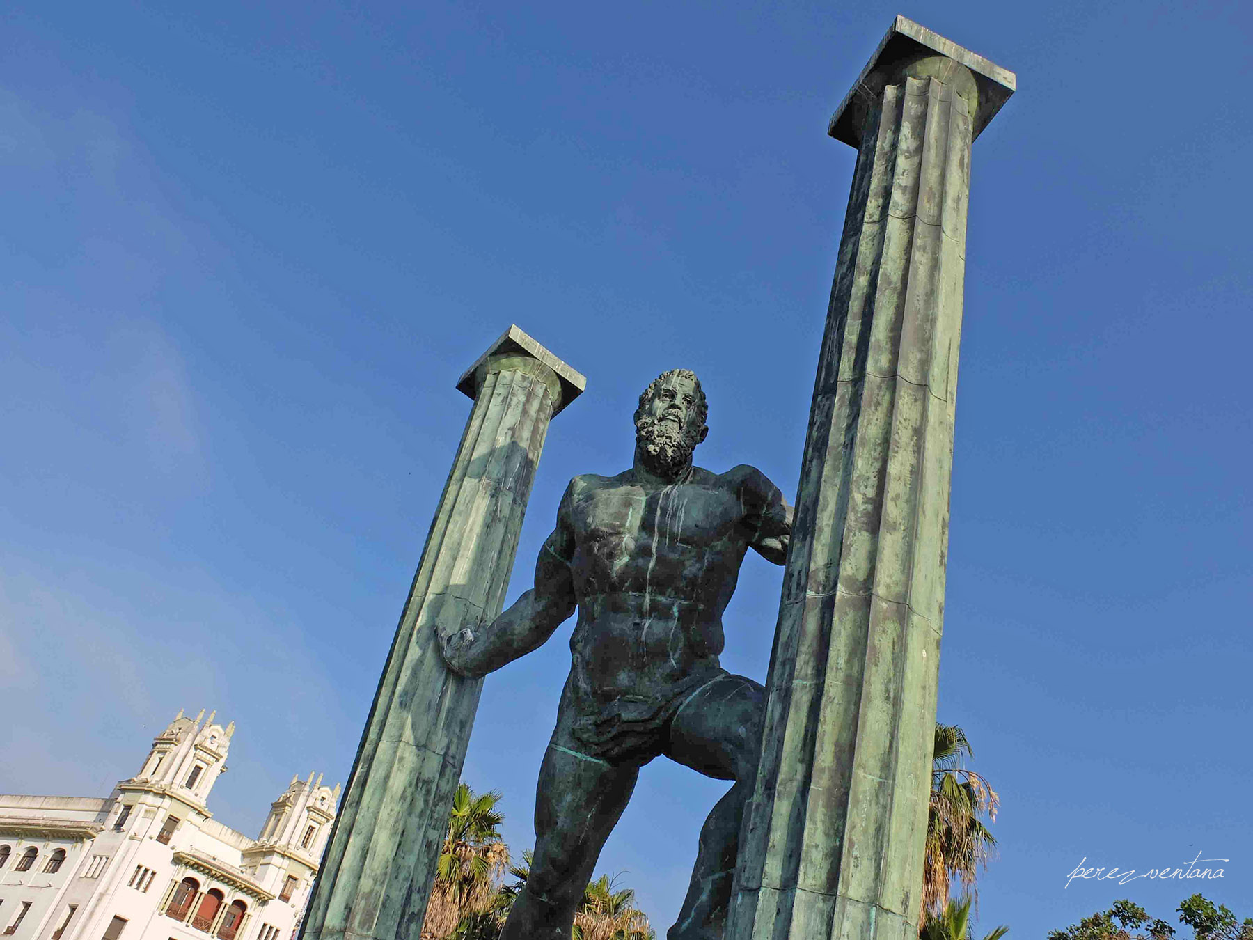Bronze statue of a muscular man between two tall, fluted columns against a clear blue sky, set in a sunny plaza with white buildings nearby.