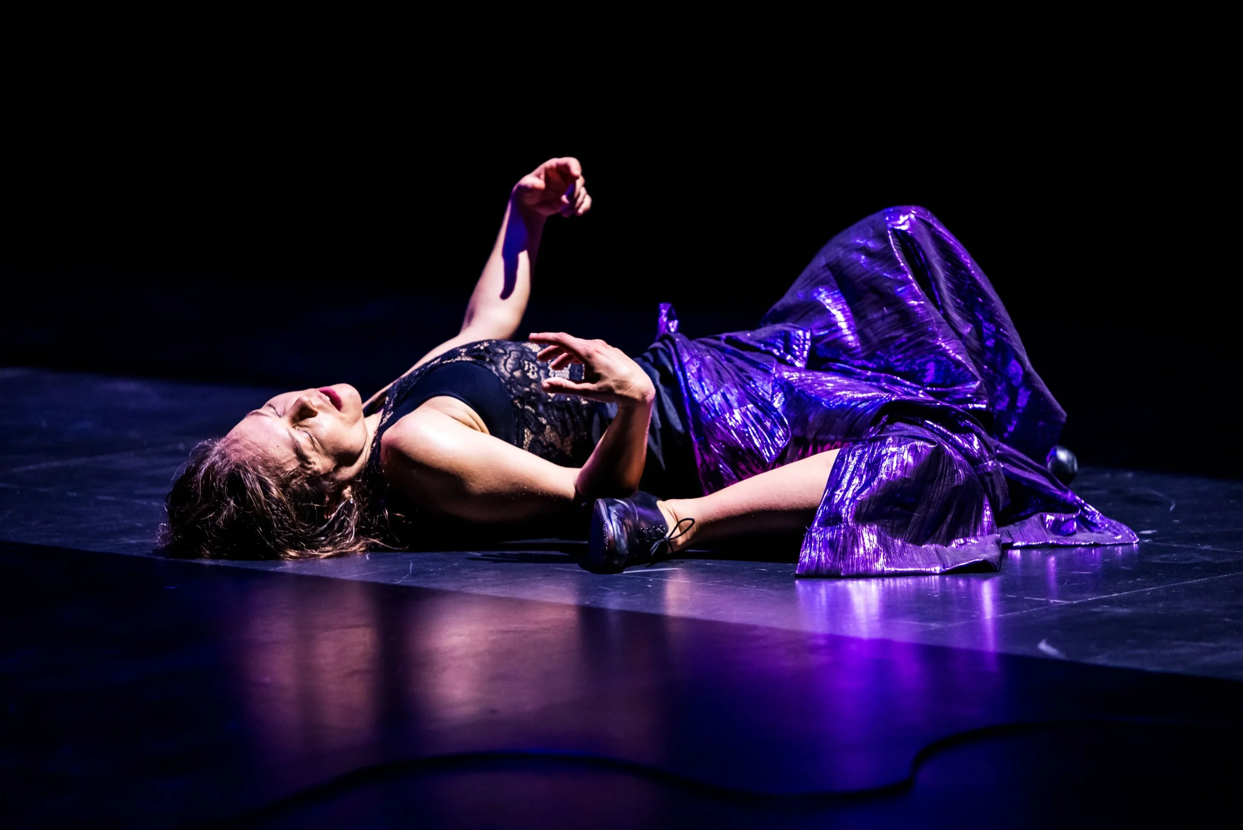 Dancer lying on a dark stage in a black lace top and shiny purple skirt, arms raised in a dramatic pose under purple light.