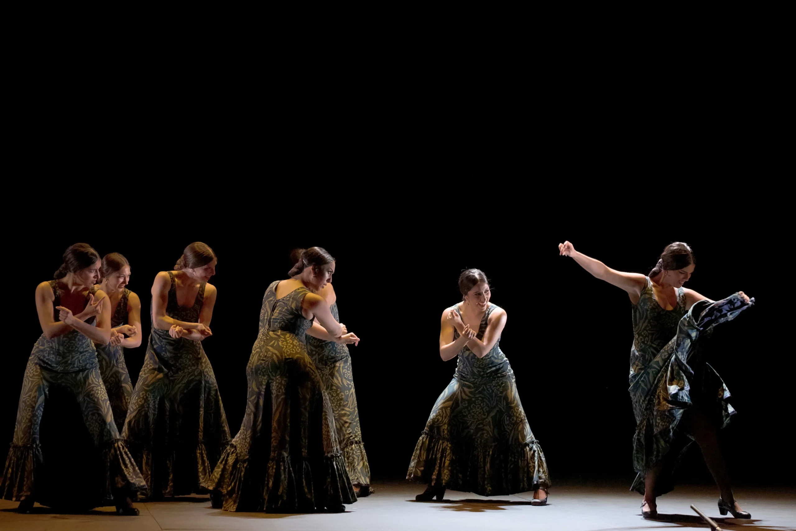 Group of female dancers in matching green patterned dresses performing a choreographed routine on a dark stage.