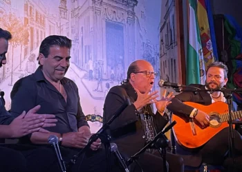 Four male musicians on a stage: one with a bright orange guitar singing into a mic while three others applaud nearby, with a mural backdrop and flags behind them at a performance venue.