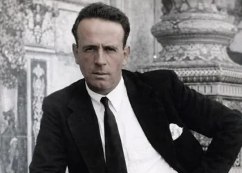 Man in a black suit and white shirt poses with hand on hip in front of an ornate stone backdrop (black-and-white photo).