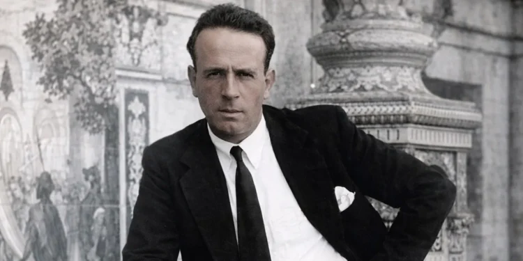 Man in a black suit and white shirt poses with hand on hip in front of an ornate stone backdrop (black-and-white photo).