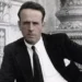 Man in a black suit and white shirt poses with hand on hip in front of an ornate stone backdrop (black-and-white photo).