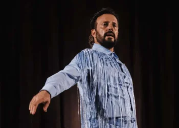 Man performing on stage in a light blue shirt with metallic fringe, arm outstretched in a dramatic pose