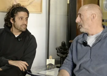 Two men sit facing each other in a casual interview setting, microphones clipped to their shirts.