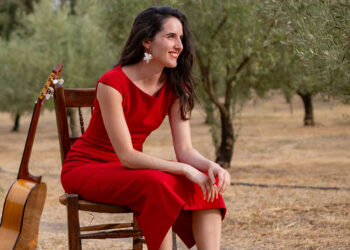 Woman in a red dress sits on a wooden chair outdoors with an acoustic guitar resting beside her in an olive grove, smiling softly.