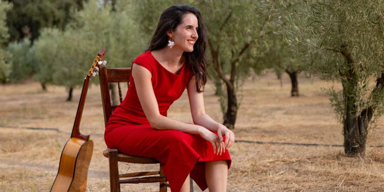 Woman in a red dress sits on a wooden chair outdoors with an acoustic guitar resting beside her in an olive grove, smiling softly.