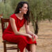 Woman in a red dress sits on a wooden chair outdoors with an acoustic guitar resting beside her in an olive grove, smiling softly.