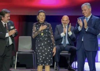 Four professionally dressed people stand on a stage, clapping; a woman in a polka-dot dress stands with a hand on her chest as red and blue lights illuminate the background.