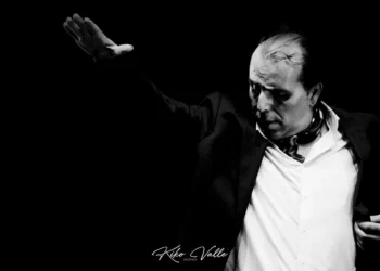 Black-and-white photo of a male performer on stage, raising one arm while looking downward, wearing a white shirt and dark jacket.