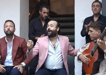 Four men performing on a small stage: center singer in a pink blazer, right-hand guitarist with an orange guitar, two others in dark jackets behind.