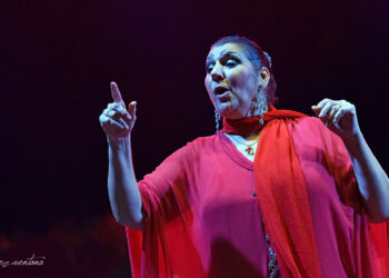 Female performer on stage singing, wearing a red shawl and pink dress, with her hands raised in expressive gesture under blue-purple lighting
