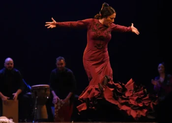 Woman in a red, ruffled dress dances on stage with arms extended, a live performance with musicians in the background.