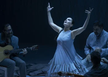 A female dancer in a light blue flowing dress raises her arms in a dramatic pose while three guitarists sit and play on a dimly lit stage.
