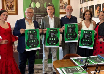 Six people pose indoors at an awards event; two women wear red flamenco dresses while others wear smart casual, each holding a green poster with circular design feature(s).