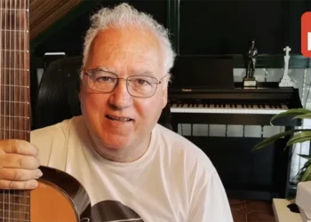 Man with gray hair and glasses holds an acoustic guitar, smiling at the camera in a home music studio with a keyboard and plants nearby, Expo Musica logo in the corner.