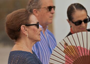 Three adults wearing sunglasses stand outdoors; the woman in the blue knit top in front holds a floral folding fan.