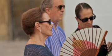 Three adults wearing sunglasses stand outdoors; the woman in the blue knit top in front holds a floral folding fan.
