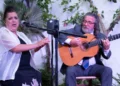 Woman in a white shawl sings into a microphone while a man in a suit plays a classical guitar outdoors among plants and trees.