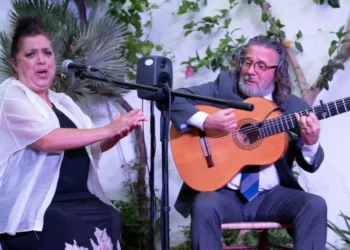 Woman in a white shawl sings into a microphone while a man in a suit plays a classical guitar outdoors among plants and trees.