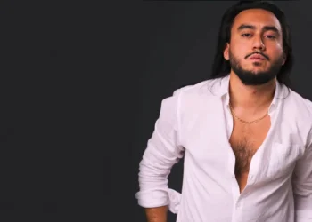 Man with long dark hair and beard wearing an open white shirt and a gold chain, posed against a dark background.