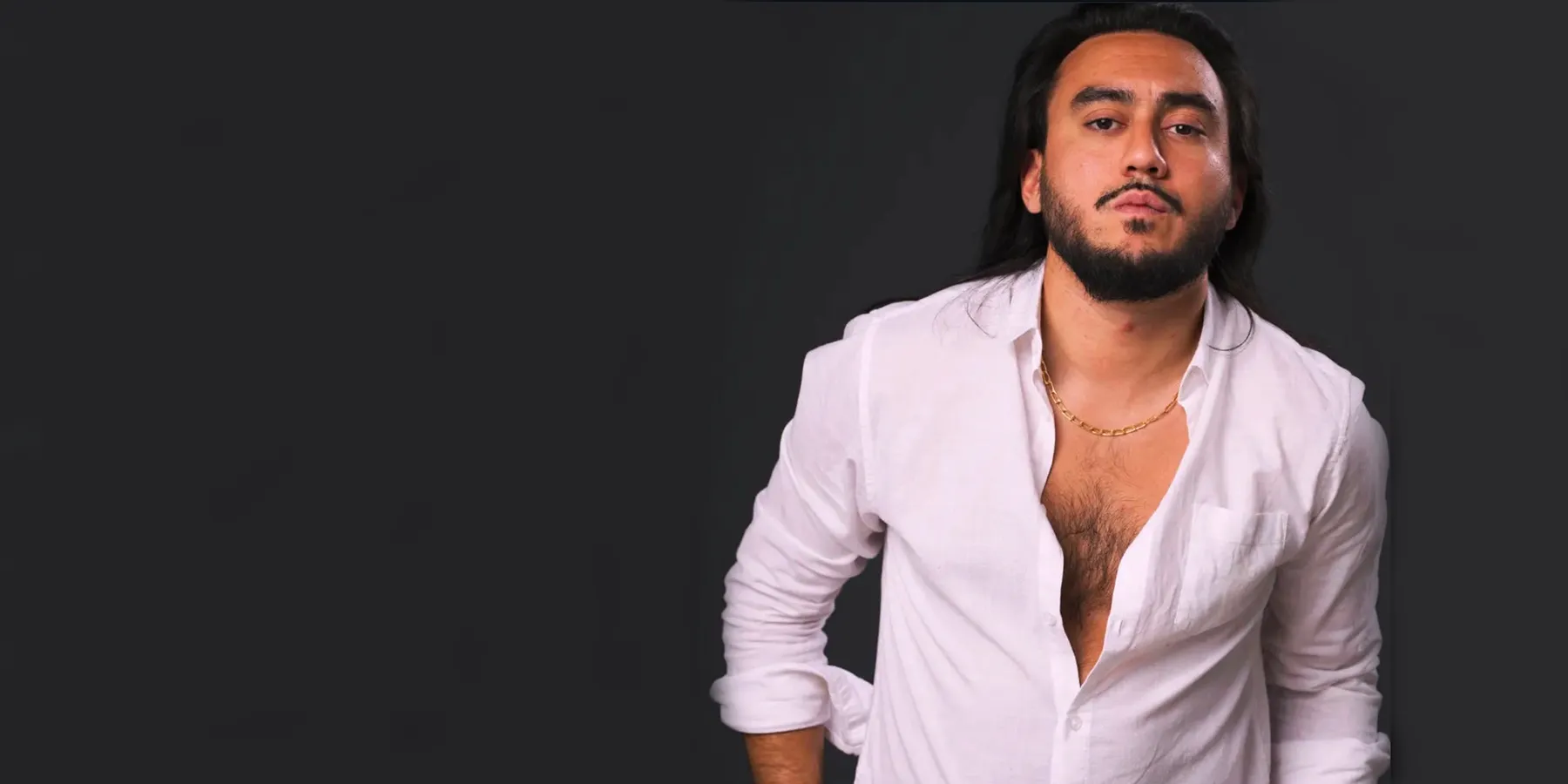 Man with long dark hair and beard wearing an open white shirt and a gold chain, posed against a dark background.