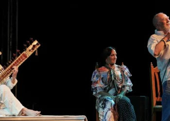 Three musicians on a dark stage: a man in a straw hat playing a sitar on the left, a woman in a colorful dress seated center, and a man singing with a microphone on the right.