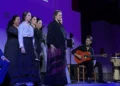 Group of flamenco performers on a purple-lit stage: a central singer in a black-purple dress, others stand behind, guitarist seated with an orange guitar, percussionist on drums nearby.