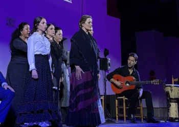 Group of flamenco performers on a purple-lit stage: a central singer in a black-purple dress, others stand behind, guitarist seated with an orange guitar, percussionist on drums nearby.