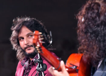 Two guitarists performing on stage; the man with shoulder-length hair and a pink shirt looks toward his fellow player as an orange guitar rests in the foreground under stage lighting.