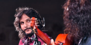 Two guitarists performing on stage; the man with shoulder-length hair and a pink shirt looks toward his fellow player as an orange guitar rests in the foreground under stage lighting.