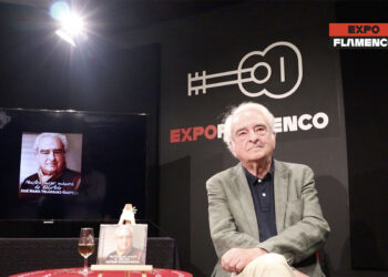 Older man in a blazer seated on stage at Expo Flamenco; a portrait poster and book sit on the table, with a screen displaying his image to the left.