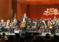 Center-stage guitarist stands amid an orchestra of string players, with the audience in the foreground and a wood-paneled concert hall behind them, during a festival performance.