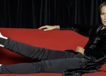 Man lounging on a bright red sofa with legs outstretched, wearing a black velvet jacket and white boots, looking at the camera.
