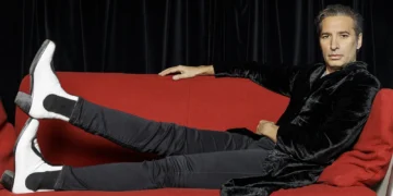 Man lounging on a bright red sofa with legs outstretched, wearing a black velvet jacket and white boots, looking at the camera.
