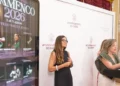 Two women at a formal event: one at a podium speaking, the other standing beside a white backdrop with Ayuntamiento de Cádiz logos.
