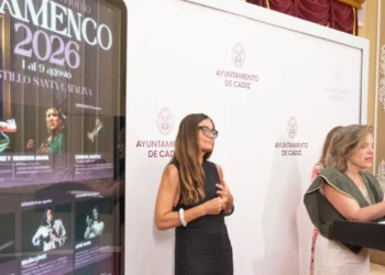 Two women at a formal event: one at a podium speaking, the other standing beside a white backdrop with Ayuntamiento de Cádiz logos.